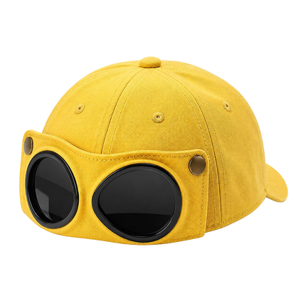 A yellow canvas baseball cap with a unique front attachment featuring two large black oval lenses and small gold-tone metal rivets, shown on a transparent checkered background.